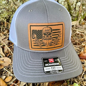 May include: Gray and white trucker hat with a brown leather patch featuring a skull with a beard and crossed wrenches. The patch also has an American flag design and the text "Garage". The hat has a Richardson 112 label.
