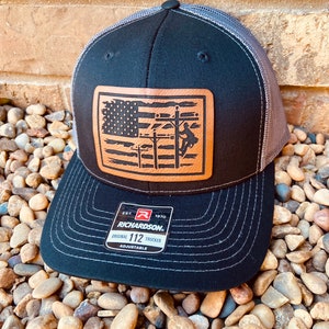 May include: Black and grey trucker hat with a brown leather patch featuring an American flag and a silhouette of a lineman working on power lines. The patch has the text "EST. 1970 RICHARDSON. ORIGINAL 112 TRUCKER ADJUSTABLE".