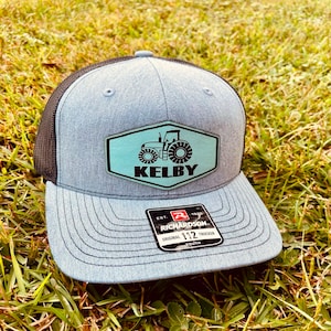 May include: A gray and black trucker hat with a green and black tractor patch that says "Kelby" on it. The hat has a black mesh back and a Richardson 112 Youth label.