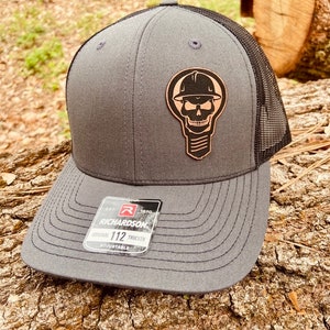 May include: A gray and black trucker hat with a black and white skull and crossbones design on the front. The hat has a black mesh back and a brown leather patch that says "EST. 1970 RICHARDSON ORIGINAL 112 TRUCKER ADJUSTABLE".