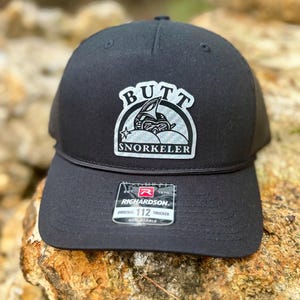 May include: Black trucker hat with a white and gray patch that says "Butt Snorkeler" in a circular design. The hat is made by Richardson 112.