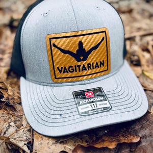 May include: A gray and black trucker hat with a gold patch that says "Vagitarian" and a carbon fiber pattern. The hat is adjustable and has a Richardson 112 label.