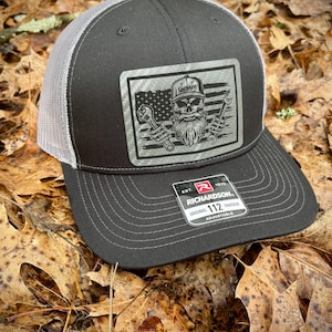May include: Black and white trucker hat with a gray mesh back. The front of the hat features a patch with a skull and crossbones, an American flag, and the word "Garage". The hat also has a Richardson 112 Trucker label.
