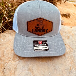 May include: Gray and white trucker hat with a brown leather patch featuring a truck and the text "LANDRY". The hat has a white mesh back and a black "Richardson 112" label.