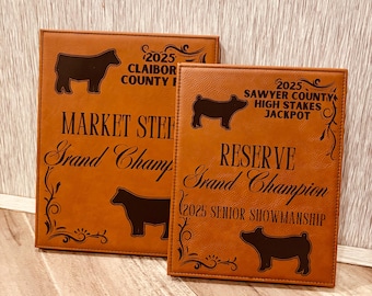 May include: Two brown leather plaques with engraved text and silhouettes of a cow and pig. The top plaque reads "2025 Claibor County Fair Market Steer Grand Champion". The bottom plaque reads "2025 Sawyer County High Stakes Jackpot Reserve Grand Champion 2025 Senior Showmanship".