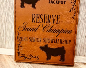 May include: A brown leather plaque with the text "2025 Sawyer County High Stakes Jackpot Reserve Grand Champion 2025 Senior Showmanship" and two black silhouettes of pigs.