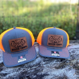 May include: Two gray and orange trucker hats with a brown leather patch that says "Bustin' Chops" and a pig head design.