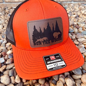 May include: Orange and black trucker hat with a brown leather patch that says "Run the Woods" and features a silhouette of trees and a bear. The hat has a black mesh back and a Richardson 112 adjustable strap.