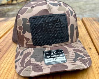 Puede incluir: Una gorra de camionero de camuflaje marrón y beige con un parche de fibra de carbono negro que presenta una torre de perforación de petróleo. La gorra tiene una parte posterior de malla negra y una etiqueta negra con el texto "EST. 1970 RICHARDSON 112 ORIGINAL TRUCKER ADJUSTABLE".