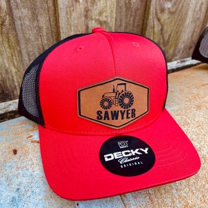 May include: Red and black trucker hat with a brown leather patch featuring a tractor and the name "SAWYER". The hat has a curved brim and a mesh back. A black and white "DECKY Classic Original" sticker is on the brim.