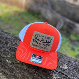 May include: Orange and white trucker hat with a brown leather patch that says "Bustin Bass & Runnin Racks". The hat is made by Richardson.