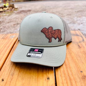 May include: A green and gray baseball cap with a brown leather patch of a steer with a floral design. The cap has the text "Richardson" and "112" on the front.