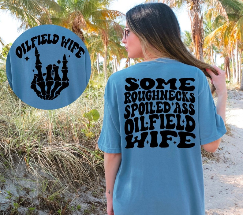 May include: A light blue t-shirt with black text that reads "Some Roughnecks Spoiled Ass Oilfield Wife" on the back. The front of the shirt has a black skeleton hand making a rock and roll sign with the text "Oilfield Wife" above it.