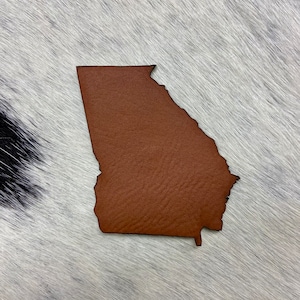 May include: A brown leather cutout of the state of Georgia. The cutout is on a white and black cowhide background.