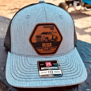 May include: A blue and black trucker hat with a Richardson patch. The hat features a brown patch with a tow truck graphic and the text "RSI TOWING & WRECKER SERVICE Tazewell, TN". The hat is labeled "Original 112 Trucker Youth".