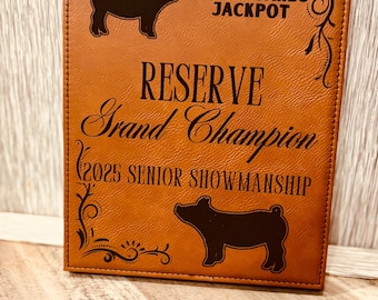 May include: A brown leather plaque with the text "2025 Sawyer County High Stakes Jackpot Reserve Grand Champion 2025 Senior Showmanship" and two black silhouettes of pigs.