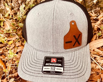 May include: Gray and black youth trucker hat with a brown leather ear tag with a black "X" on it. The hat has a black mesh back and a Richardson 112 Original Trucker label.