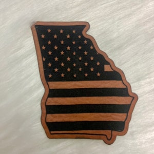 May include: A brown leather cutout of the state of Georgia with the American flag design. The stars are white and the stripes are red and white.