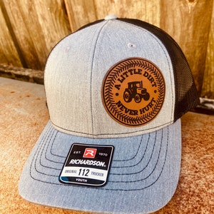 May include: A gray and black youth trucker hat with a brown leather patch that says "A Little Dirt Never Hurt" with a tractor silhouette. The hat has a Richardson 112 label.