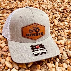May include: Gray and white trucker hat with a brown leather patch featuring a tractor and the word "DENVER". The patch also has the text "EST. 1970 RICHARDSON. ORIGINAL 112 TRUCKER YOUTH".