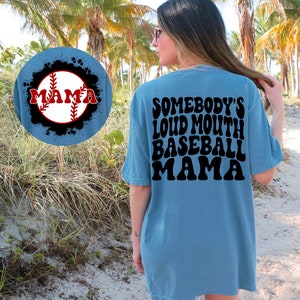 May include: A light blue t-shirt with the text "Somebody's Loud Mouth Baseball Mama" in black retro font on the back. The front of the shirt features a baseball with the word "Mama" in red letters on a blue background.