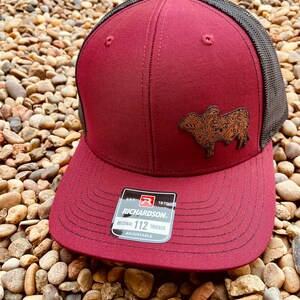 May include: A maroon and black trucker hat with a brown leather patch featuring a stylized cow design. The hat has an adjustable strap with the text "Est. 1970 Richardson Original 112 Trucker Adjustable".