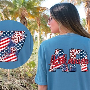 May include: A blue t-shirt featuring a red, white and blue American flag design in the shape of the state of Arkansas. The letters "AR" are also on the shirt, with the same design.
