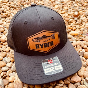 May include: Black trucker hat with a brown leather patch featuring a fish and the word "RYDER". The hat has a mesh back and a Richardson label with the text "EST. 1970 RICHARDSON. ORIGINAL 112 TRUCKER YOUTH".