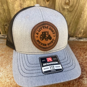 May include: A gray and black trucker hat with a brown leather patch that says "A Little Dirt Never Hurt" with a tractor silhouette. The hat is a youth size and has the Richardson logo and "Original 112 Trucker Youth" text on the label.