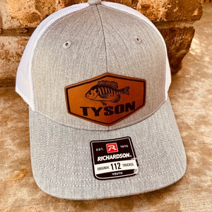 May include: Gray and white youth trucker hat with a brown leather patch featuring a black and white fish and the text "TYSON". The hat also has a white label with the text "EST. 1970 RICHARDSON. ORIGINAL 112 TRUCKER. YOUTH".