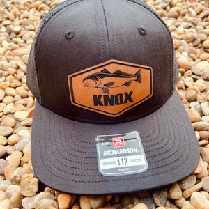 May include: Black and brown youth trucker hat with a leather patch featuring a fish and the word "KNOX". The hat is made by Richardson and has the text "EST. 1970 ORIGINAL 112 TRUCKER YOUTH" on the back.