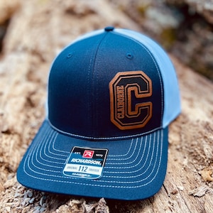 May include: A navy blue and light blue trucker hat with a brown leather patch featuring a large "C" with the word "CLAIBORNE" above it. The hat has a Richardson 112 adjustable strap.
