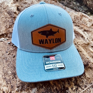May include: A light blue youth trucker hat with a brown leather patch featuring a shark and the text "WAYLON". The hat has a white mesh back and a black label with the text "RICHARDSON. ORIGINAL 112 TRUCKER YOUTH" and a red "R" logo.