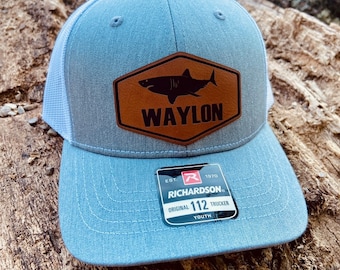 May include: A light blue youth trucker hat with a brown leather patch featuring a shark and the text "WAYLON". The hat has a white mesh back and a black label with the text "RICHARDSON. ORIGINAL 112 TRUCKER YOUTH" and a red "R" logo.