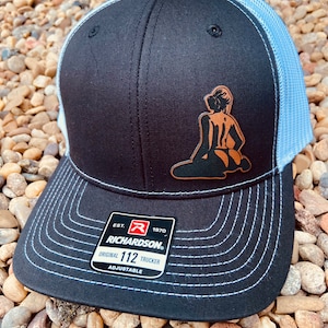 May include: Black trucker hat with a light blue mesh back and a brown leather patch featuring a silhouette of a woman. The hat has a curved brim with white stitching and an adjustable strap. The Richardson brand label is visible.