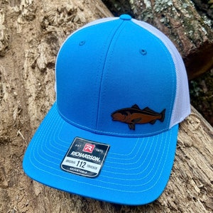 May include: A light blue trucker hat with a white mesh back. The hat has a brown leather fish patch on the front. The hat is adjustable and has the Richardson 112 logo on the side.