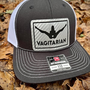May include: A black and white trucker hat with a white mesh back. The hat has a carbon fiber-like patch with a silhouette of a person and the word "VAGITARIAN" in black letters. The hat also has a red and white label that says "Richardson 112 Original Trucker Adjustable".
