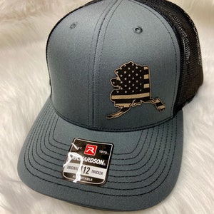 May include: A gray and black trucker hat with a gold outline of the state of Alaska with an American flag design. The hat has an adjustable strap with the Richardson logo and "Original 112 Trucker" text.