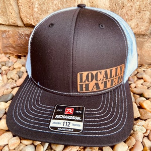 May include: Black and white trucker hat with a brown patch that says "Locally Hated". The hat has a blue mesh back and a Richardson 112 label.