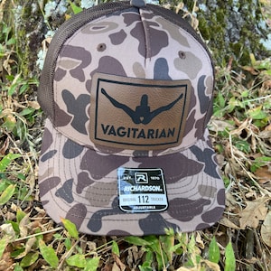 May include: A brown and tan camouflage trucker hat with a brown leather patch that says "Vagitarian" and a black mesh back. The hat has a black adjustable strap with the text "Est. 1970 Richardson Original 112 Trucker Adjustable".