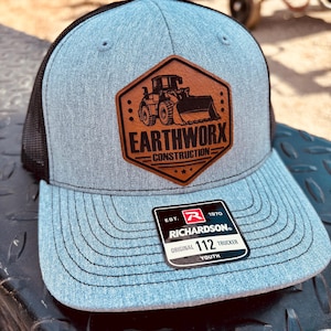 May include: A gray and black trucker hat with a Richardson 112 Youth tag. The hat features a brown leather patch with the text "EARTHWORX CONSTRUCTION" and a graphic of a front-end loader.