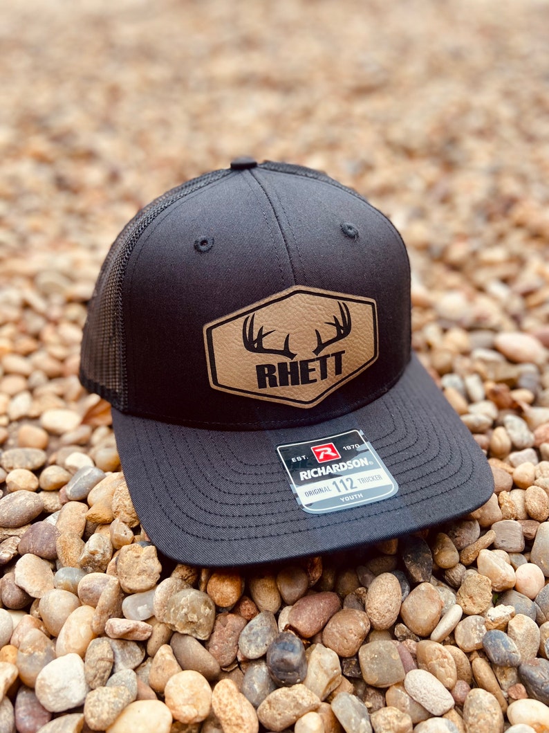 May include: Black mesh trucker hat with a brown leather patch featuring a buck's antlers and the word "Rhett". The hat is labeled "Richardson 112 Youth".