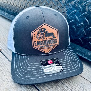 May include: A black and white trucker hat with a leather patch featuring the text "EARTHWORX CONSTRUCTION" and a bulldozer graphic. The hat has a curved brim and a mesh back. The hat is adjustable.