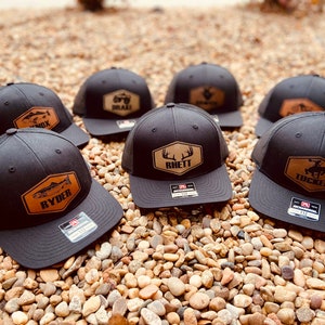 May include: Six black trucker hats with brown leather patches featuring various designs and names, including "Ryder", "Rhett", and "Tucker".