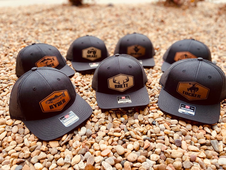 May include: Six black trucker hats with brown leather patches featuring various designs and names, including "Ryder", "Rhett", and "Tucker".
