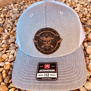May include: A gray trucker hat with a white mesh back and a leather patch featuring a skull with crossed axes and the letters "FD". The hat has a curved brim and the brand tag "RICHARDSON 112 TRUCKER".