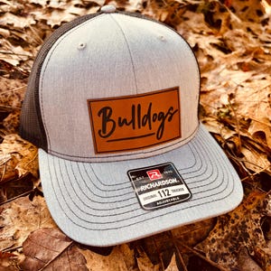 May include: A gray and black trucker hat with a brown leather patch that says "Bulldogs" in white script. The hat is adjustable and has a black mesh back.