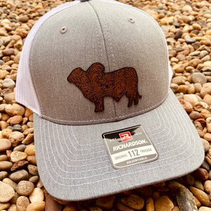 May include: Gray and white trucker hat with a brown leather patch of a steer with a floral design. The patch is on the front of the hat. The hat has a white mesh back and a Richardson 112 label on the side.