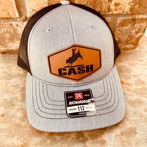 May include: Gray and black youth trucker hat with a brown leather patch that says "CASH" and a bull rider silhouette. The patch has a stitched border. The hat has a Richardson 112 label.