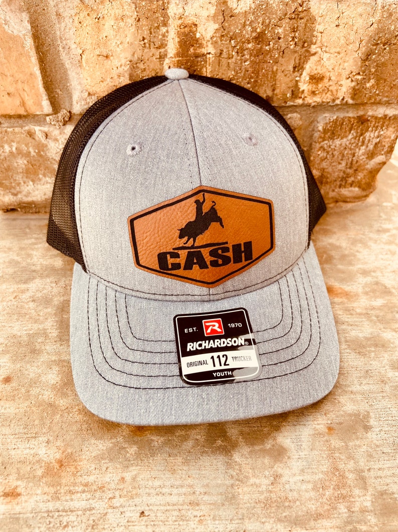 May include: Gray and black youth trucker hat with a brown leather patch that says "CASH" and a bull rider silhouette. The patch has a stitched border. The hat has a Richardson 112 label.